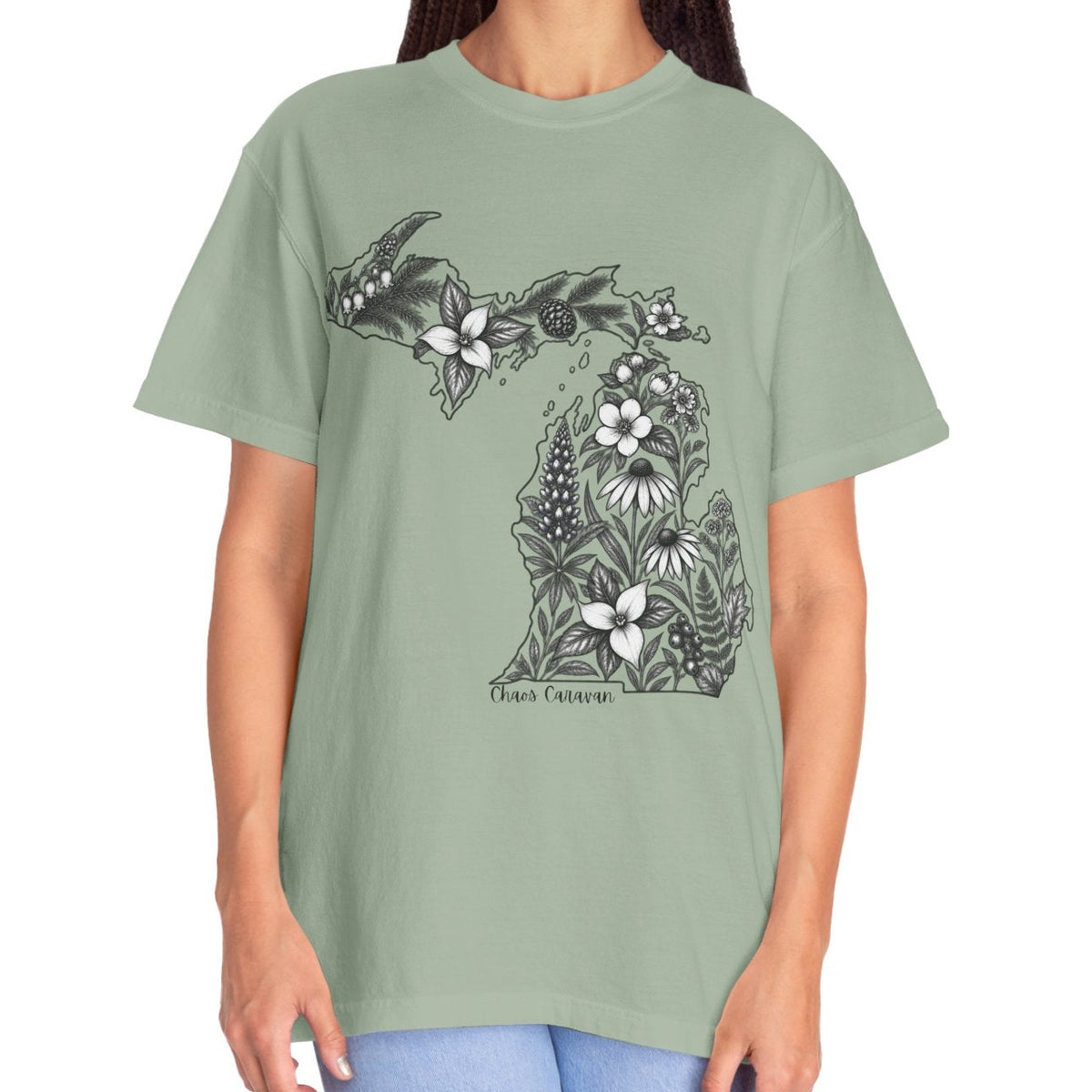 A woman modeling a sage green t-shirt with a black and white floral Michigan map graphic on the front, paired with light blue jeans, against a plain white background.