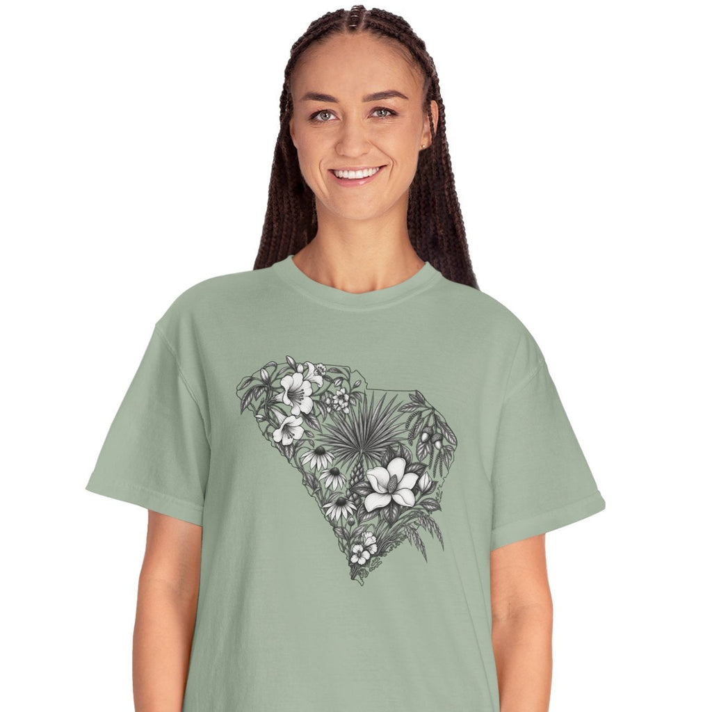 A woman modeling a sage green short-sleeve t-shirt with a black and white tropical floral heart graphic on the chest, paired with light blue jeans, against a plain white background.