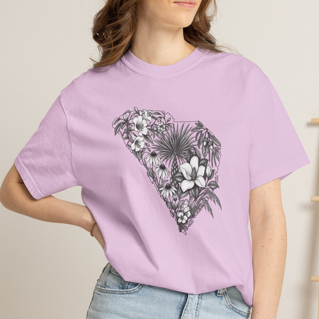 A woman wearing a light purple short-sleeve t-shirt with a black and white tropical floral graphic in the shape of South Carolina, paired with light blue denim shorts, standing in a brightly lit room.
