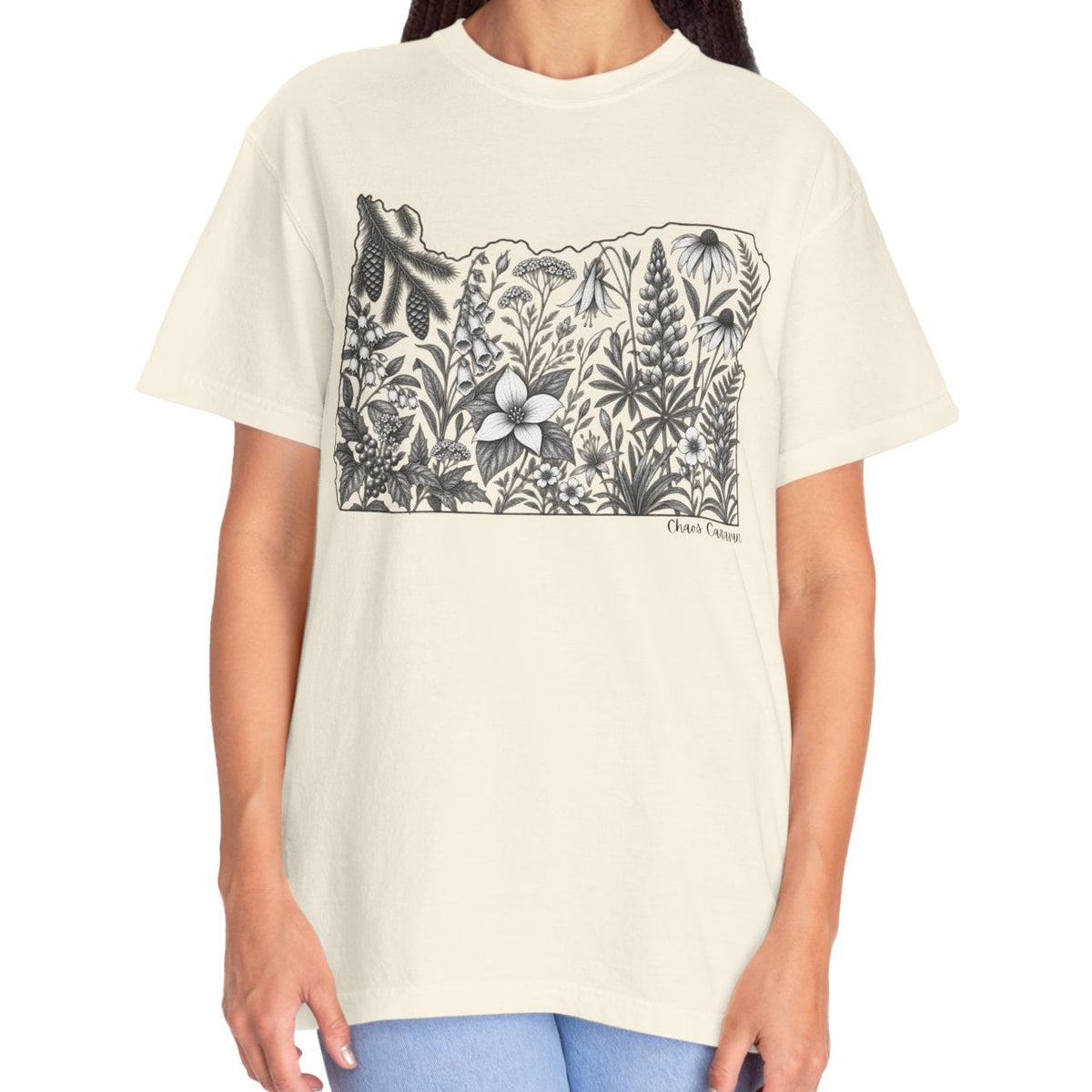 A woman modeling a cream-colored short-sleeve t-shirt with a black line art graphic of the state of Oregon filled with wildflowers and a wolf silhouette. She is wearing light blue jeans against a solid white background.