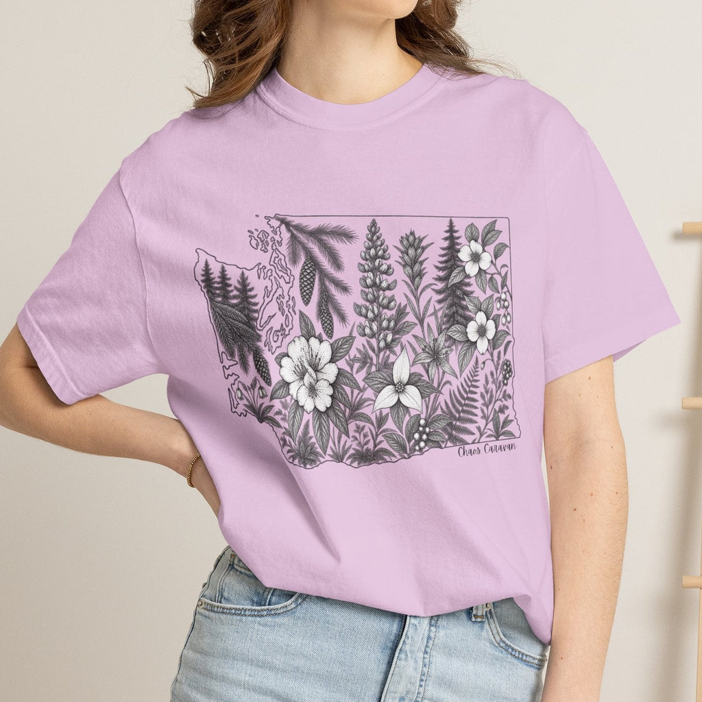 A woman wearing a light purple short-sleeve graphic t-shirt with a black and white botanical design of ferns and flowers, paired with light blue denim shorts, standing in a brightly lit room.