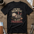 A black graphic t-shirt laid flat on a rustic wooden table, surrounded by lifestyle props including a coffee mug, notebooks, and silverware. The t-shirt features a humorous graphic of an opossum rolling over with the text 'I'M NOT DRAMATIC I'M JUST THEATRICAL.