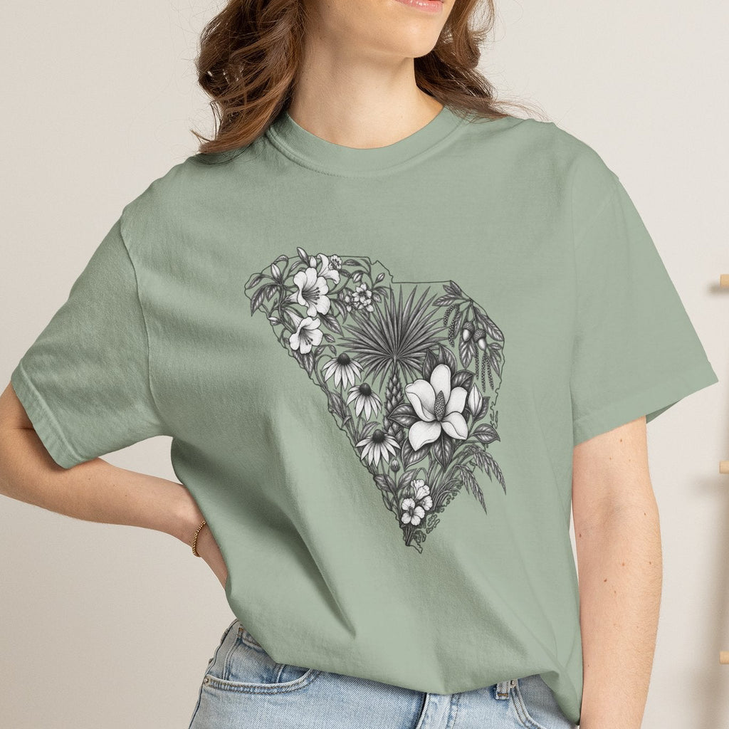 A woman wearing a sage green graphic t-shirt with a black and white floral design shaped like the state of South Carolina, paired with light blue denim shorts in a cozy indoor setting.
