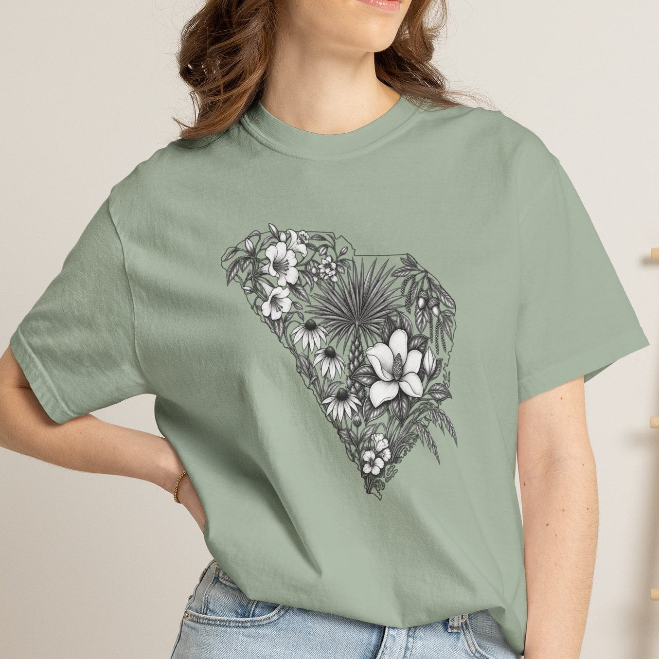 A woman wearing a sage green graphic t-shirt with a black and white floral design shaped like the state of South Carolina, paired with light blue denim shorts in a cozy indoor setting.