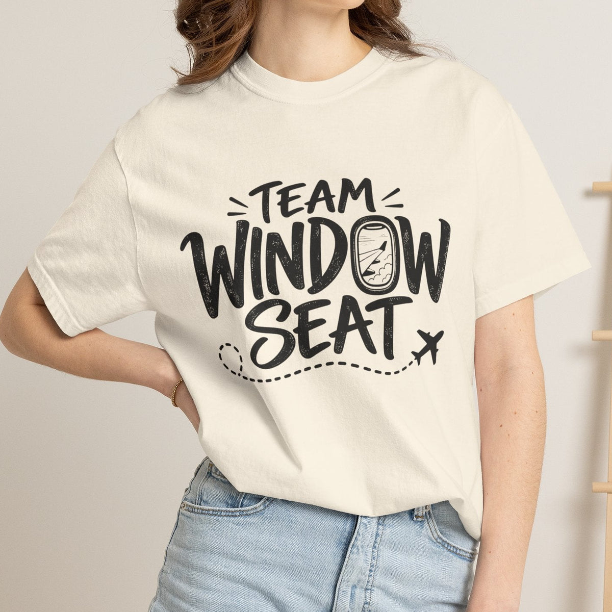 A woman wearing a cream-colored graphic t-shirt with the text 'TEAM WINDOW SEAT' in black, paired with light-wash denim shorts, standing in a brightly lit room.