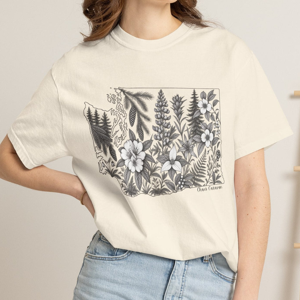 A woman wearing a cream-colored short-sleeve t-shirt with a black and white botanical graphic of pine trees and wildflowers inside the state outline of Washington. She is also wearing light-wash denim shorts, standing in a brightly lit room with a wooden ladder and woven furniture in the background.