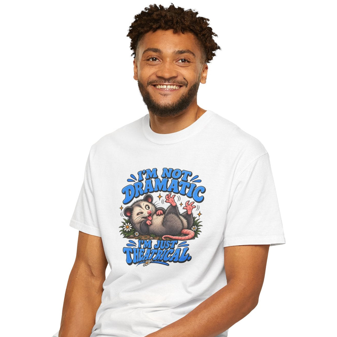 A man sitting on a black stool wearing a white graphic t-shirt and light blue jeans against a plain white background. The t-shirt features a colorful cartoon sloth graphic with the text 'I'M NOT DRAMATIC, I'M JUST THEATRICAL'.