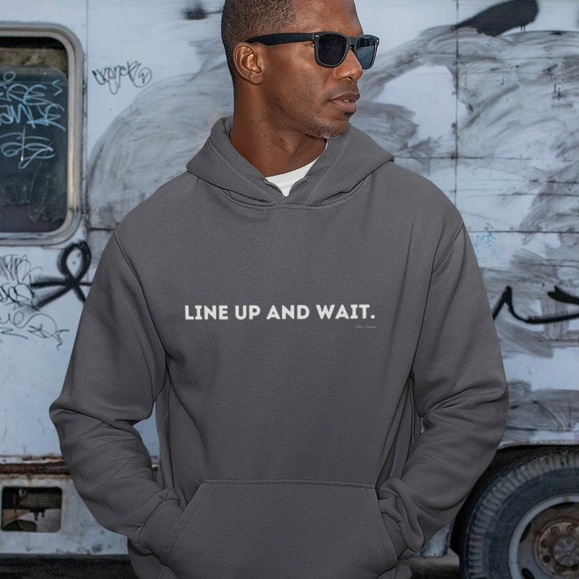 Man wearing a gray hoodie with 'LINE UP AND WAIT.' text, standing in front of a graffiti-covered wall.